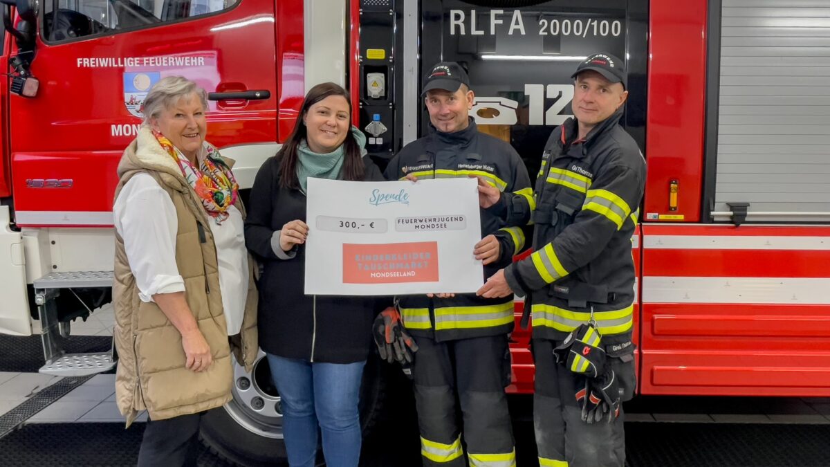 Vier Personen stehen vor einem Feuerwehrauto. Zwei Personen überreichen symbolisch eine Spende an den Feuerwehrkommandanten und seinen Stellvertreter. Die Szene strahlt Engagement und Unterstützung für die Feuerwehr aus, während die Spende übergeben wird.