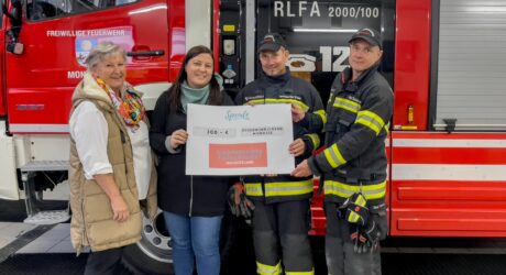 Vier Personen stehen vor einem Feuerwehrauto. Zwei Personen überreichen symbolisch eine Spende an den Feuerwehrkommandanten und seinen Stellvertreter. Die Szene strahlt Engagement und Unterstützung für die Feuerwehr aus, während die Spende übergeben wird.