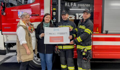 Vier Personen stehen vor einem Feuerwehrauto. Zwei Personen überreichen symbolisch eine Spende an den Feuerwehrkommandanten und seinen Stellvertreter. Die Szene strahlt Engagement und Unterstützung für die Feuerwehr aus, während die Spende übergeben wird.
